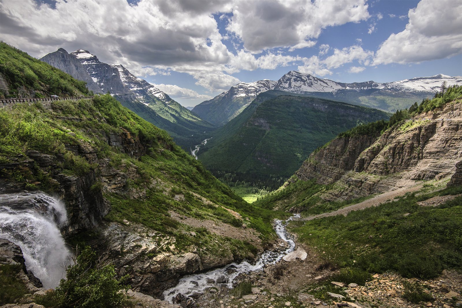 Going-to-the-Sun Road follows a green mountainside above a waterfall and deep valley in Glacier National Park.